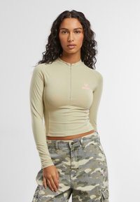 Lys grønn langermet zip-top med en tettsittende design og en liten rosa logo. Kombinert med kamuflasje cargo-bukser med sidelommer.