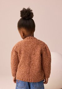 Tekturowy brązowy sweter dzianinowy z grubymi rękawami, prążkowanym dołem i okrągłym dekoltem, noszony na niebieskich dżinsach z haftem kwiatowym.