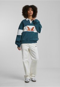 Hanorac supradimensionat teal cu dungi albe și bleumarin, prezentând un grafic cu creaturi portocalii cu aripi. Asortat cu pantaloni albi cu croi larg.