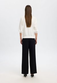 Weißer Pullover mit Puffärmeln, gerippten Bündchen und Saum, kombiniert mit schwarzen weitem Hose und schwarzen Absatzschuhen. Glatter Stofftextur.