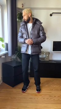 Moški z blond lasmi, oblečen v sivo puhasto jakno, belo srajco, črne hlače in športne copate, stoji v prostoru s rastlinami in šivalnim strojem.