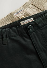 Kaksi paria housuja, yksi musta ja yksi beige. Molemmissa on nappikiinnitys ja vyötäröllä "NEXT SUPPLY" -merkinnällä varustetut tarrat.
