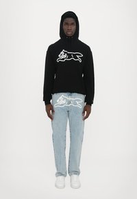 Hanorac negru cu grafică albă a unui animal, asociat cu blugi deschiși la culoare având un design similar. Modelul poartă teniși albi; fundal simplu.