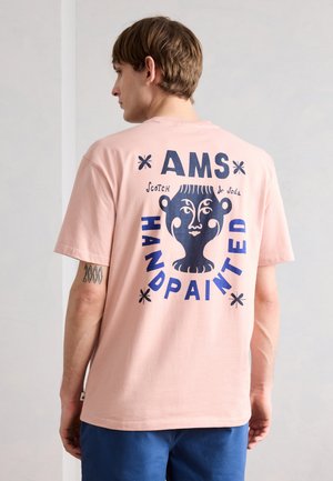 Ung mand vendt væk iført en pink T-shirt med et blåt abstrakt ansigt og teksten "AMS HANDPAINTED Scotch & Soda" på ryggen.