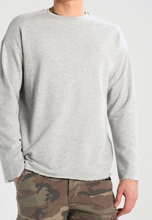 Mężczyzna ubrany w jasnoszarą koszulę z długim rękawem i spodnie bojówki w kamuflażu, stojący z rękami swobodnie opuszczonymi wzdłuż ciała.