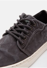 Szürke vászon sportcipő, amely textúrázott felülettel, fehér gumitalppal és kerek fűzőkkel rendelkezik, amelyek fém fűzőlyukakon át vezetnek. Zöld logóval díszítve.