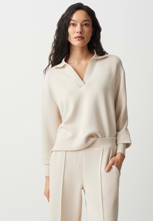 Femme aux longs cheveux foncés portant un pull crème à col en V et un pantalon plissé assorti, une main dans la poche, fond neutre.