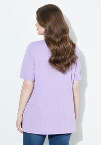 Mujer con cabello largo y ondulado castaño, vistiendo una camisa de manga corta lila y jeans azules, de pie contra un fondo blanco, mirando hacia la izquierda.