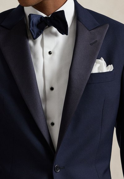 Man wearing dark blue tuxedo with satin lapels, white dress shirt, black studs, black bow tie, and white pocket square.