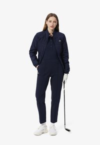 Tenue de golf marine comprenant une veste zippée et un pantalon ajusté assorti, fabriqués dans un tissu texturé. Accentué par un logo vert.