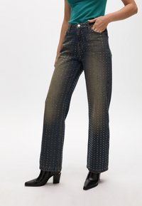 Dark blue denim jeans with golden rivets, straight-leg cut, high waist, and side pockets, paired with black heeled ankle boots.