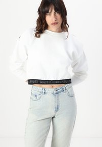 Sweatshirt branco cropped com uma gola larga, apresentando uma cintura elástica com a marca em preto. Calças de ganga ligeiramente desbotadas de cintura alta em azul claro.
