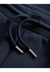 Cuerdas de tiro en azul marino con tejido texturizado y puntas metálicas en pantalones deportivos. Presentan una superficie suave y una cinturilla elástica.