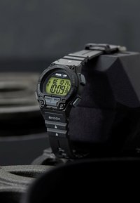 Black digital watch with a rectangular face, green LCD display, rubber strap, and features for timekeeping, lap timing, and recall functions.