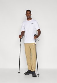 Λευκό μπλουζάκι με μικρό μαύρο λογότυπο, καφέ παντελόνι cargo με φερμουάρ και μαύρα παπούτσια πεζοπορίας. Το άτομο κρατάει μπαστούνια πεζοπορίας.