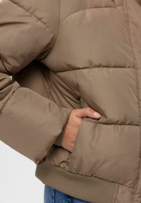 Jakna s punjenjem u boji khaki, s podstavljenom teksturom, rebrastim rubom i bočnom džepom; glatka tkanina s opuštenim krojem.