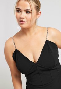 Schwarzes Kleid mit tiefem V-Ausschnitt, ausgestattet mit glänzenden, kristallverzierten Trägern. Glatte Textur und figurbetontes Design mit gefälteltem Detail.