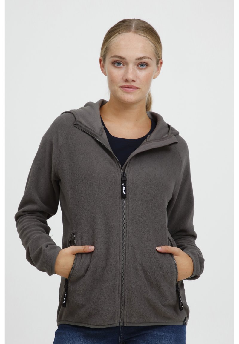 Chaqueta de forro polar gris con capucha, cremallera frontal, bolsillos laterales y puños de canalé. Textura suave y diseño ajustado. El modelo está de pie con las manos en los bolsillos.