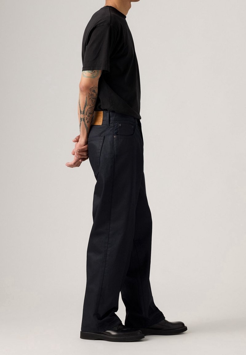 Homme en t-shirt noir et pantalon noir à jambes larges avec des bras tatoués, debout les mains jointes derrière le dos sur un fond uni.