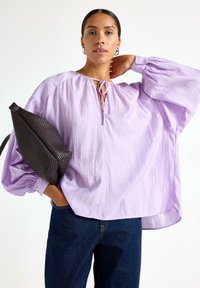 Blusa lavanda con maniche a palloncino, tessuto testurizzato e scollo con laccetto. Abbinata a jeans scuri e una borsa intrecciata scura.