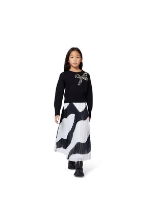 Pulôver de malha preto com embellishments de joias no peito, combinado com uma saia plissada em padrão de vaca em preto e branco e botas pretas.