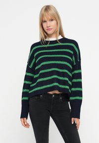 Joven mujer con cabello rubio lacio que lleva un suéter de punto a rayas azul marino y verde y jeans negros, de pie frente a un fondo blanco.