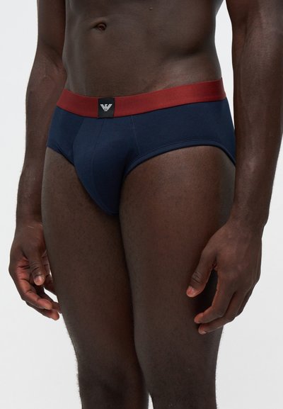 Mannequin masculin portant un slip bleu marine avec une ceinture rouge et un petit patch logo noir au centre devant.