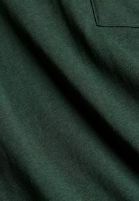 Tissu vert foncé avec des plis doux visibles et un coin de poche cousue en haut à droite.