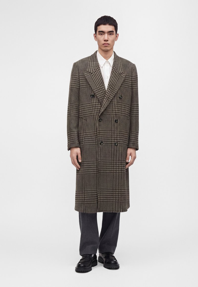 Homme debout portant un long manteau croisé à carreaux marron et noir sur une chemise blanche, un pantalon gris et des chaussures noires sur fond blanc.