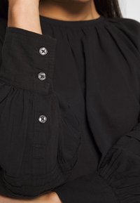 Svart långärmad topp med rynkad tyg vid ärmarna och silverknappar vid manschetterna. Mjuk, strukturerad väv med en avslappnad passform.