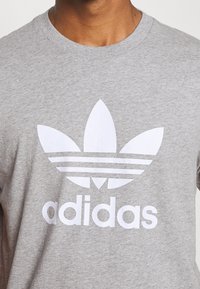 Šedé bavlněné tričko s velkým bílým logem Adidas trefoil a třemi vodorovnými pruhy nad názvem značky. Standardní design s kulatým výstřihem.