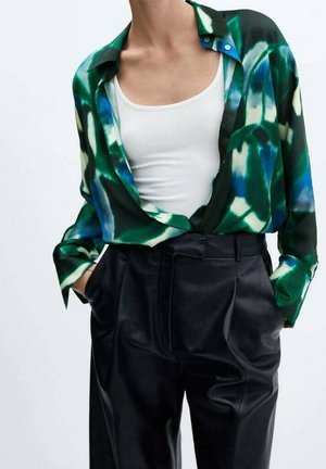 Personne portant une chemise ouverte à motifs verts et bleus sur un débardeur blanc, associée à un pantalon noir taille haute, mains dans les poches.