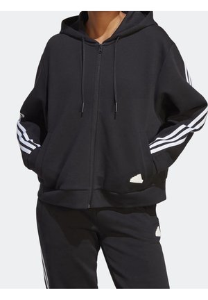 Personne portant un sweat à capuche noir zippé et un pantalon assorti avec des bandes blanches Adidas sur les manches et les côtés du pantalon.