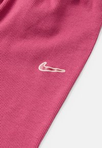 Roza rebrasta tkanina s belim Nike logotipom. Tekstura je gladka z navpičnimi črtami, kar ji daje prilegan videz.