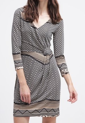 Femme portant une robe portefeuille à motifs noir et blanc avec des manches trois-quarts et des motifs géométriques en bordure.