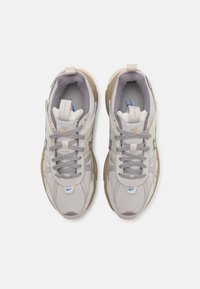 Ljust grå atletiska skor med strukturerad yta, krämfärgade detaljer och en brun gummisula; har en vadderad tunga och blå logodetalj.