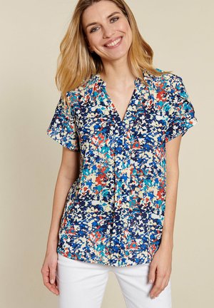 Mujer sonriente con cabello rubio lleva una blusa floral multicolor de manga corta y pantalones blancos sobre un fondo beige.