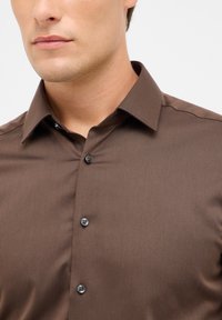 Braune Hemdbluse aus strukturiertem Stoff mit einem Punktkragen und vier dunklen Knöpfen auf der Vorderseite. Einfaches, tailliertes Design.