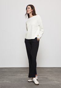 Kvinna med brunt hår iklädd en vit texturerad tröja med lång ärm, svarta raka byxor och krämfärgade sneakers, som står med händerna i fickorna.