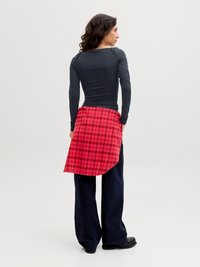 Kvinne med langt, mørkt, krøllete hår, iført en tettsittende svart genser med lange ermer, en rød rutete skjorte knyttet rundt livet, vide mørke bukser og svarte sko.