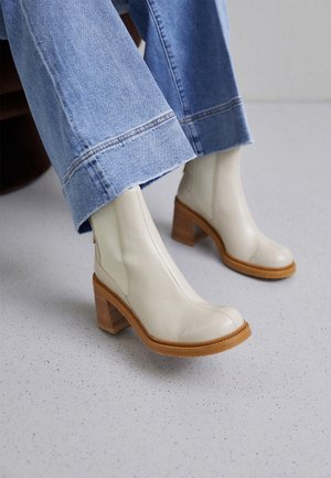 Białe skórzane botki z kwadratowym noskiem i brązowym, warstwowym obcasem. Połączone z jasnoniebieskimi szerokimi spodniami, ustawione na posadzce w plamki.