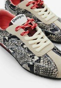 Sneaker mit Schlangenledermuster, in Beige- und Schwarztönen, ausgestattet mit pinken Schnürsenkeln, strukturiertem Material und einem weißen Markenlabel auf der Zunge.