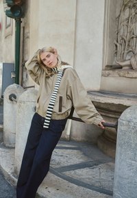 Veste beige à fermeture éclair avec une capuche, ornée d'accents brun foncé et d'une sangle rayée. Pantalon ample bleu foncé, posant devant un mur en pierre.