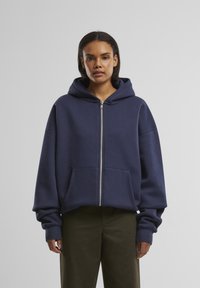 Marineblå zip-up hoodie lavet af blødt stof med en stor hætte, drop shoulder og frontlommer; har ribbede manchetter og afslappet pasform.