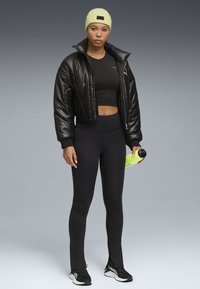 Musta puuvillane jope, istuv must crop top, kõrge vöökoht mustad leggingsid, neoonroheline beanie, hoiab käes erksat roheline veepudelit.