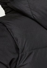 Černá polstrovaná bunda s texturovanou tkaninou, vybavená kapucí a viditelnými stehy. Materiál vypadá lehce a voděodolně.