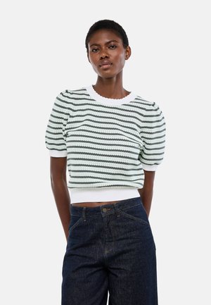 Femme portant un haut en maille à manches courtes rayé blanc et vert avec des manches bouffantes et un jean taille haute bleu foncé, debout devant un fond blanc.