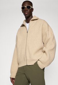 Maglione beige a coste con zip e collo alto, dalla vestibilità oversize e tessuto testurizzato, abbinato a pantaloni verde oliva e occhiali da sole chiari.