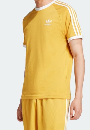 Personne portant un t-shirt Adidas jaune moutarde et un short assorti avec des bandes blanches sur les manches et les côtés, debout devant un fond uni.