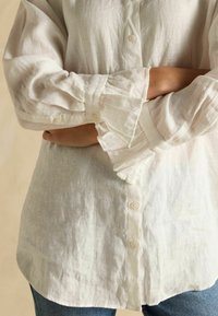 White linen shirt with buttoned front and ruffled cuffs, featuring a relaxed fit and a slightly curved hem. Visible texture of fabric.